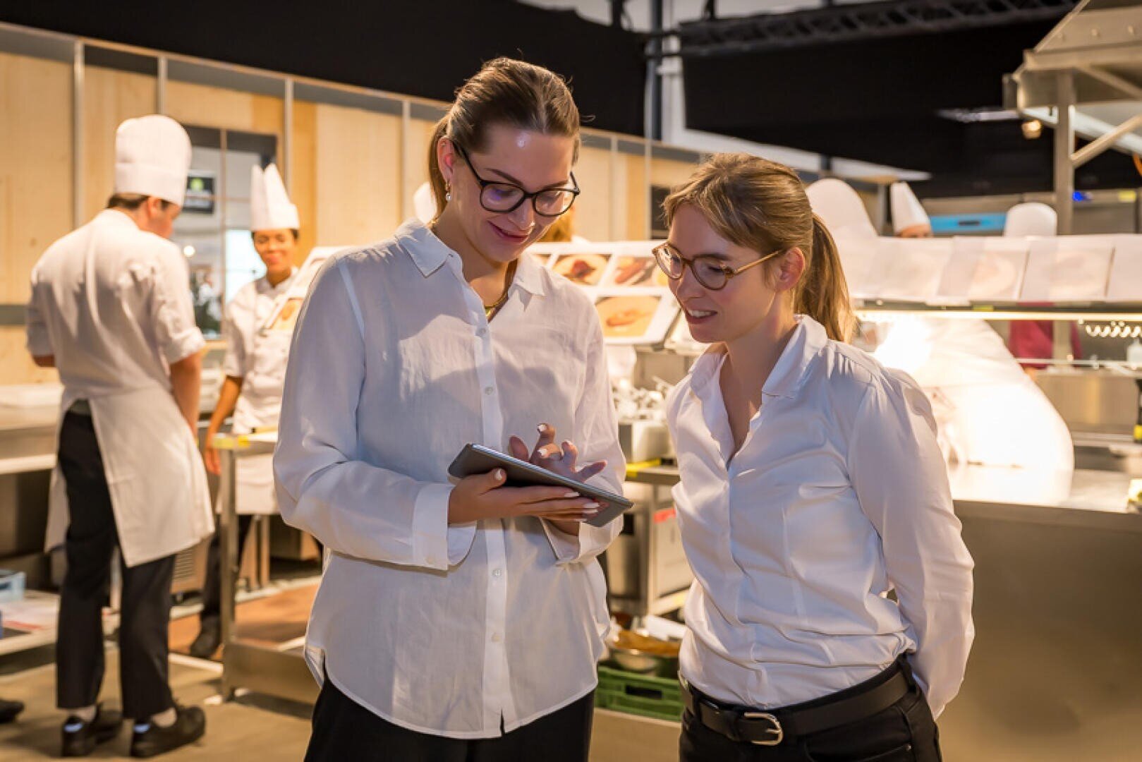 Zwei Frauen in weißen Hemden stehen in einer Profiküche und schauen auf ein Tablet. Im Hintergrund sind Köche in weißen Uniformen und Kochmützen bei der Arbeit. Die Atmosphäre ist hell und geschäftig.