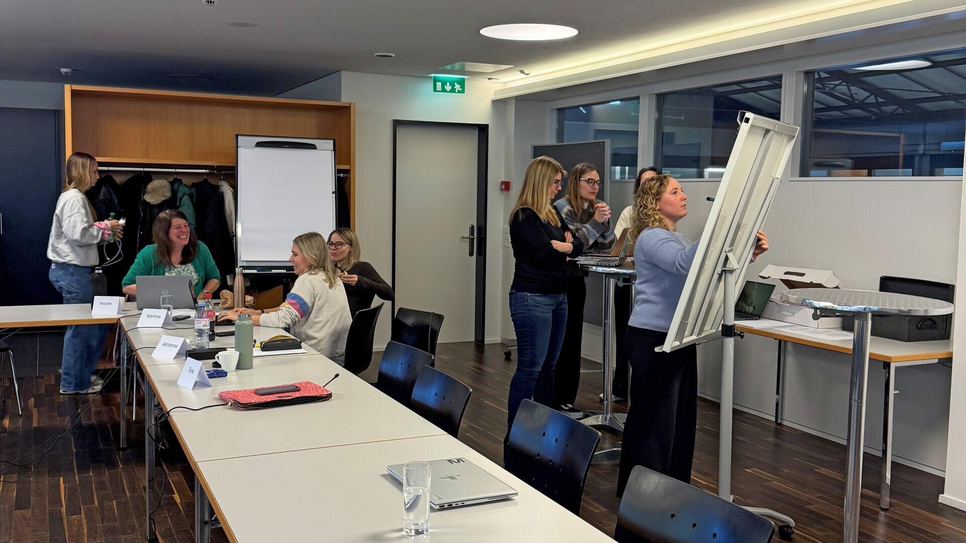 Eine Gruppe von Frauen arbeitet in einem modernen Besprechungsraum zusammen. Einige sitzen an Tischen mit Laptops, während andere an einem Flipchart stehen und diskutieren. Die Atmosphäre scheint produktiv und engagiert zu sein.