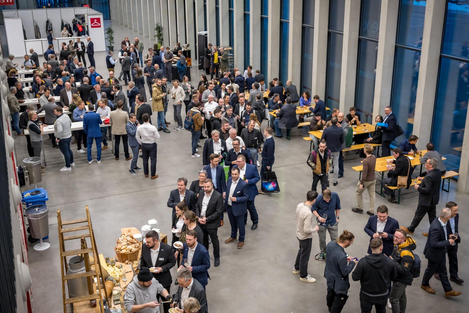 Eine große Gruppe von Menschen in Geschäftskleidung mischt sich unter die Leute, knüpft Kontakte und isst in einer geräumigen, modernen Halle mit hohen Fenstern. Einige versammeln sich in der Nähe der Tische mit den Speisen, während andere sich in kleinen Gruppen unterhalten oder an Holztischen sitzen.
