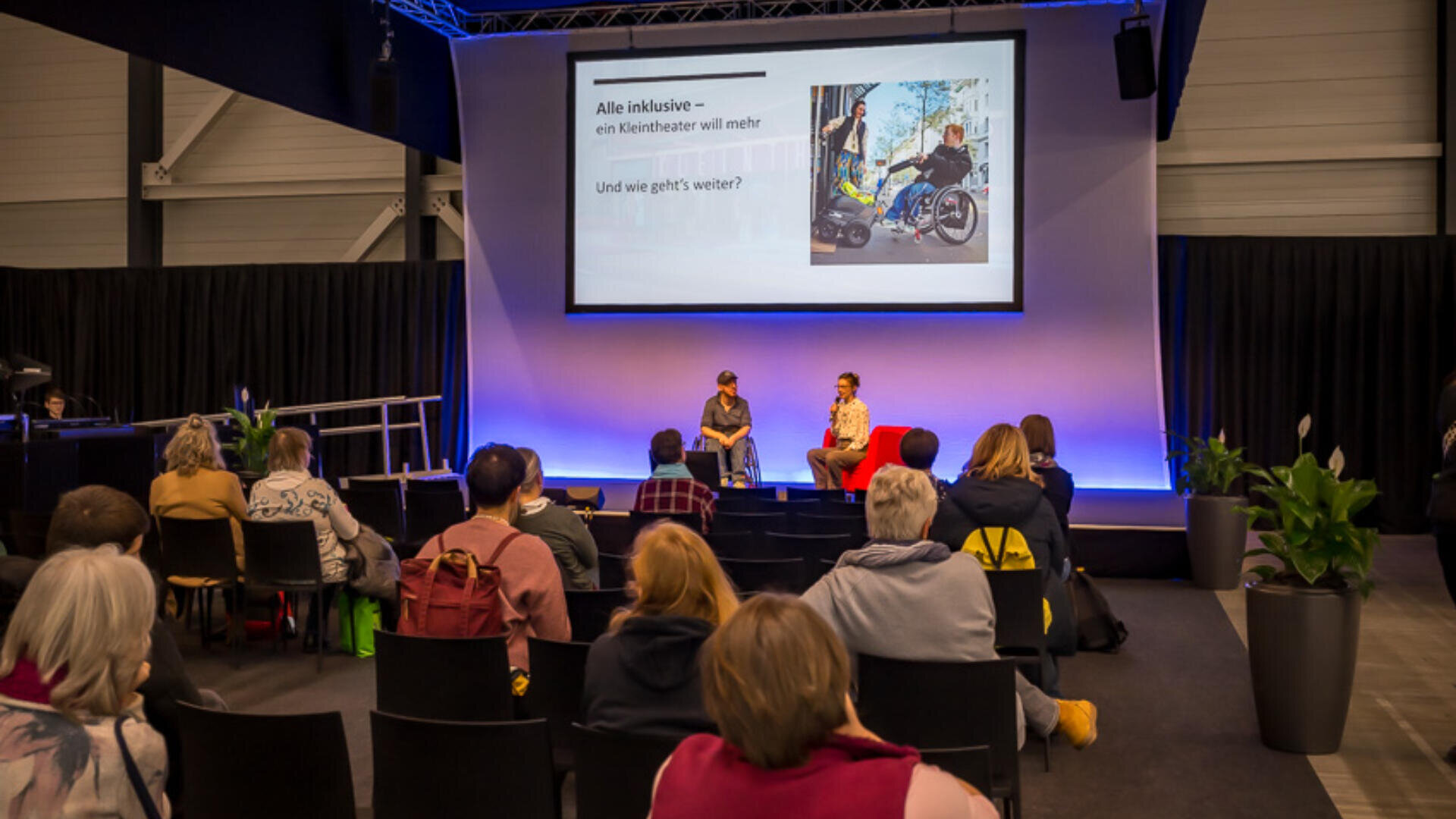 Zwei Personen sitzen auf der Bühne und diskutieren vor Publikum. Auf einem großen Bildschirm hinter ihnen sind ein Foto und ein deutscher Text zu sehen. Der Raum ist modern, blau beleuchtet und verfügt über mehrere Sitzreihen.
