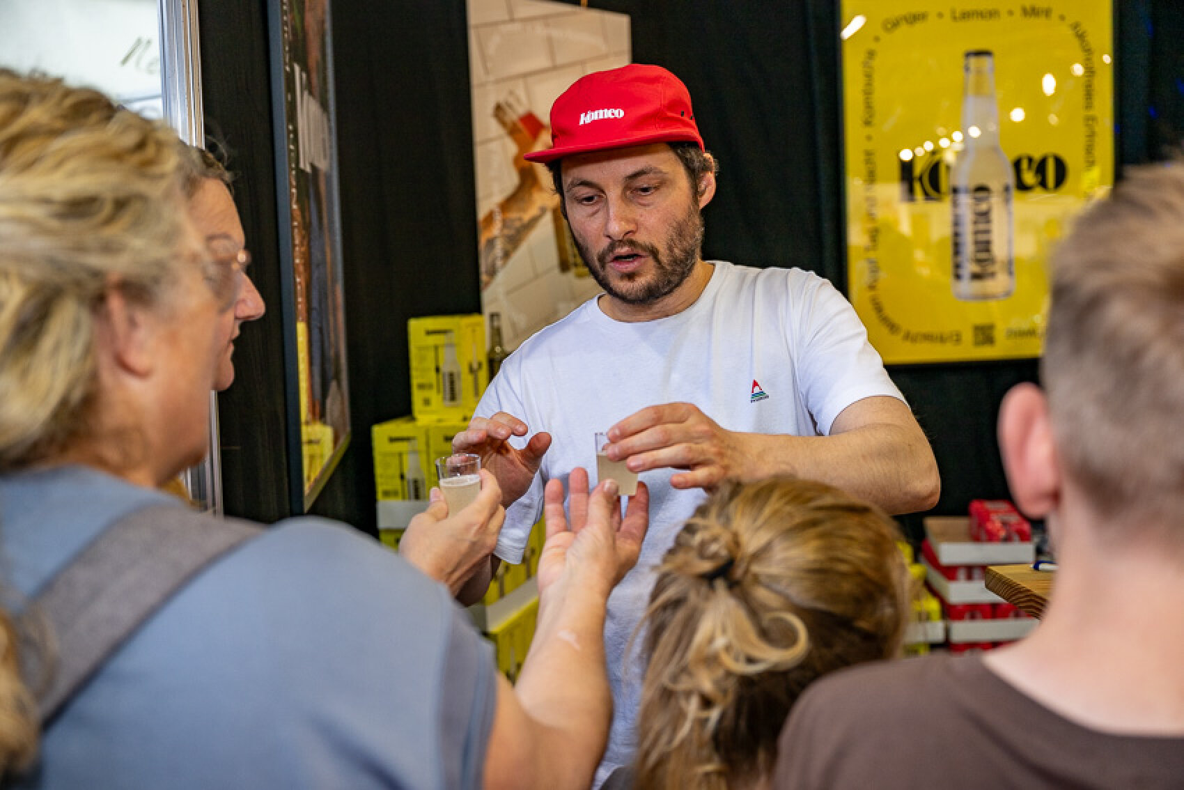 Ein Mann mit roter Mütze reicht einer Frau an einem Stand einen kleinen Probierbecher, während zwei Kinder und ein weiterer Erwachsener zuschauen. Im Hintergrund ist ein gelbes Plakat mit einer Flasche zu sehen.