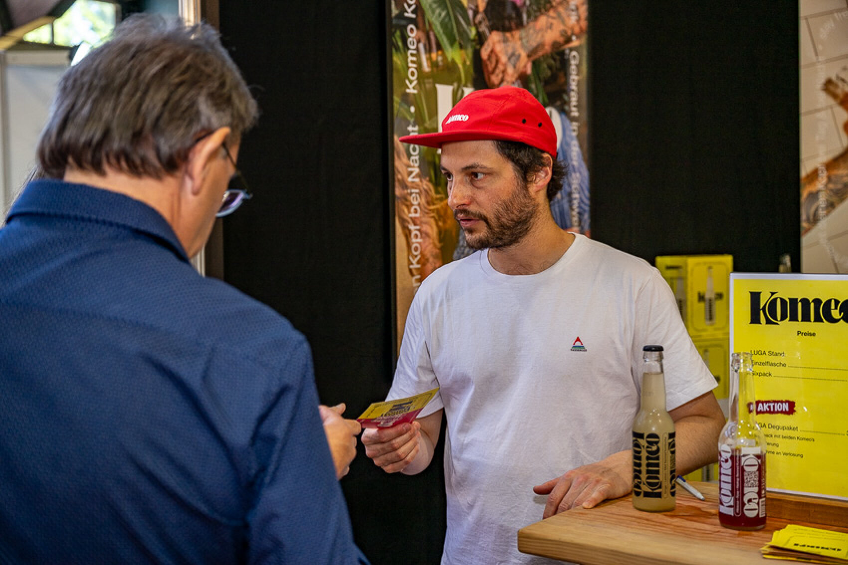 Ein Mann in weißem T-Shirt und roter Mütze steht hinter einem Tresen und unterhält sich mit einem anderen Mann, der einen Flyer in der Hand hält. Vor ihm stehen Flaschen mit der Aufschrift „Komeco“, im Hintergrund sind Werbeplakate zu sehen.